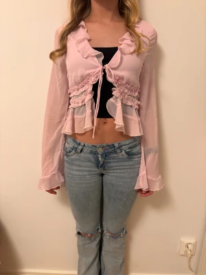 Ljusrosa omlott blus med volanger - Supersöt ljusrosa blus med omlott och knyt framtill. Blusen har långa, fladdriga ärmar och volangdetaljer längs halsringning och midja. Materialet är tunt och transparent, vilket ger en luftig känsla. Aldrig använd förutom för foton. Modellen är ungefär 158 cm och bär storlek S