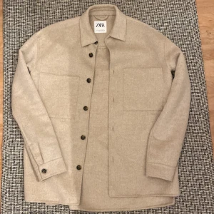 Beige overshirt från Zara, S - Snygg beige overshirt från Zara i storlek S. Jackan har klassisk krage, stora bröstfickor och knäppning med mörka knappar framtill. Materialet är mjukt och känns som ull, perfekt för lager-på-lager. Stilren och enkel design som funkar till många outfits.
