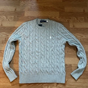 Stickad Ralph Lauren tröja  - Snygg grå kabelstickad tröja från Polo Ralph Lauren med klassisk rund hals och broderad vit logga på bröstet. Tröjan har ribbade muddar och är långärmad, perfekt för dig som gillar stilren och tidlös design. Helt ny, priset är ej hugget i sten så var inte rädda att ställa frågor 😁