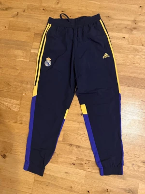 Real Madrid Adidas träningsbyxor blå/gul - Snygga träningsbyxor från Adidas med Real Madrid-logga. Byxorna är marinblå med gula och lila detaljer längs benen, elastisk midja och muddar vid benslut. Perfekta för fotboll eller chill, med klassiska Adidas-ränder på sidorna.