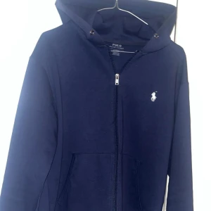 hoodie från Polo Ralph Lauren - Snygg marinblå hoodie från Polo Ralph Lauren med klassisk vit logga på bröstet. Tröjan har huva, dragkedja framtill och två fickor. Perfekt för chill dagar och enkel att matcha med jeans eller joggers. Storlek S. Hoodin har inte snören, därav priset! ✌️❤️