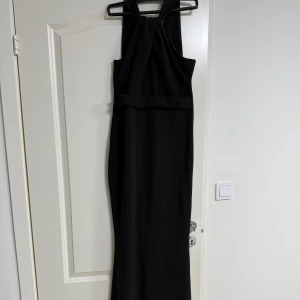 Svart långklänning med omlott - Svart långklänning med snygg omlott-detalj framtill och breda axelband. Klänningen har en elegant, figurnära passform och är ärmlös. Materialet känns mjukt och har ett fint fall, perfekt för dig som gillar stilrena och klassiska looks. Goddiva klänning från bubbleroom. 