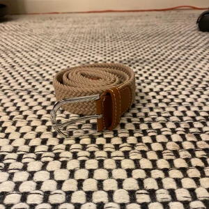 Beige flätat bälte med skinn-detaljer - Snyggt beige flätat bälte med bruna skinn-detaljer och silvrigt spänne. Perfekt accessoar för att lyfta din outfit med en stilren och klassisk touch. Bältet har en mjuk och flexibel känsla tack vare det stickade materialet. 100cm långt