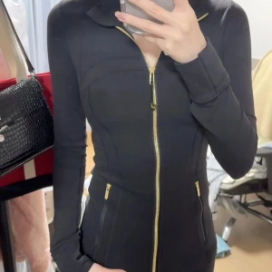 Svart slim träningsjacka med guldzip - Slimmad svart jacka med snygga guldiga dragkedjor framtill och på fickorna. Långärmad och figurnära passform, perfekt för dig som gillar sportig och stilren look. Materialet känns mjukt och stretchigt, vilket gör den bekväm att bära både till och från gymmet.