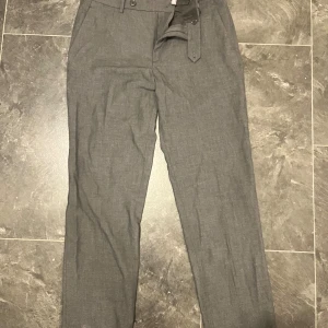 Grå kostymbyxor  - Snygga grå kostymbyxor med raka ben och tillhörande bälte. Byxorna har hällor, knäppning fram och två bakfickor med knapp. Perfekta för en clean och stilren look. Materialet känns lätt och passar bra till skjorta eller t-shirt.