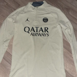 Beige PSG långärmad fotbollströja Jordan - Säljer en beige långärmad PSG tröja från Jordan med snyggt mönster i tyget, halv dragkedja och svart Paris Saint-Germain-logga på bröstet. Qatar Airways tryck på framsidan och Jordan-logga. Slim fit i ventilerande material.