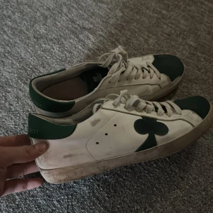 Vita och gröna sneakers The Gallerist - Snygga vita sneakers från The Gallerist med gröna detaljer och klövertryck på sidan och hälen. Skorna har snörning, platt sula och rund tå. Materialet är skinn och de har en cool kontrast mellan vitt och mörkgrönt. Perfekta för dig som gillar unika sneakers med grafiska inslag.