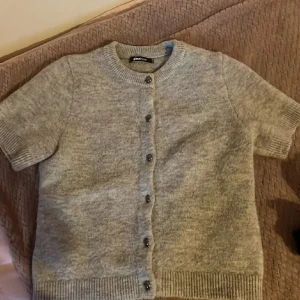 Grå kortärmad kofta från Gina Tricot - Supermysig grå kofta från Gina Tricot med korta ärmar och rund hals. Koftan är stickad och har snygga knappar framtill samt ribbade muddar vid ärmslut och nederkant. 