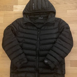 Svart dunjacka från Arc'teryx - Säljer en svart dunjacka från Arc'teryx med huva och snygga horisontella sömmar. Jackan har dragkedja framtill och logga på ärmen samt huvan. Perfekt för kalla dagar.