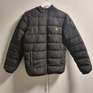 Svart pufferjacka från Lyle & Scott - Svart pufferjacka från Lyle & Scott med huva och gul logga på bröstet. Jackan har horisontella quiltade sömmar, dragkedja framtill och två sidofickor. Materialet är syntet och jackan har en normal passform, perfekt för kalla dagar.