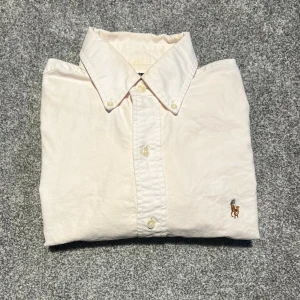 Ljusrosa  skjorta från Polo Ralph Lauren - Ljusrosa skjorta från Polo Ralph Lauren i slim fit med klassisk krage och knappar framtill. Broderad logga med polospelare på bröstet. Tillverkad i mjuk bomull, perfekt för dig som gillar stilrena och tidlösa plagg.