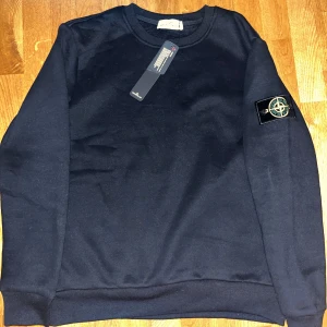 Svart sweatshirt från Stone Island - Svart sweatshirt från Stone Island med klassisk rund hals och ribbade muddar. Tröjan har lång ärm och den ikoniska Stone Island-patchen på vänster ärm. Materialet är mjuk bomull som känns skön mot huden. Perfekt för dig som gillar stilrena och snygga streetwear-plagg. Lite stor i storlek.