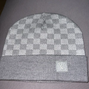 Grå rutig Louis Vuitton mössa - Snygg grå mössa från Louis Vuitton med klassiskt rutigt mönster och LV-logga framtill. Mössan har uppvikt kant och är stickad i mjukt material, perfekt för kyliga dagar.