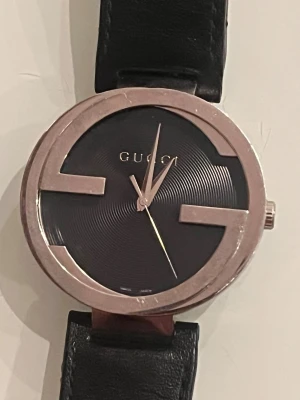 Gucci svart klocka med läderband - Säljer en stilren limited edition Gucci-klocka med rund boett i silverfärgad metall och svart urtavla med subtila cirkelmönster. Klockan har ett svart läderband med klassiskt spänne och insida i beige med präglat Gucci-mönster. Perfekt för dig som gillar exklusiv design. Behov av batteribyte
