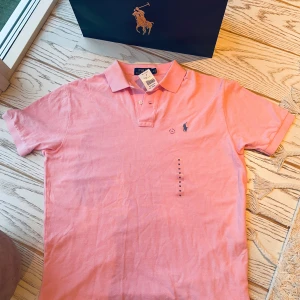 Rosa pikétröja från Polo Ralph Lauren - Snygg rosa pikétröja från Polo Ralph Lauren med klassisk krage och korta ärmar. Tröjan har den ikoniska broderade loggan på bröstet och är tillverkad i mjuk bomull. Perfekt för dig som gillar stilren och sportig look. Helt ny 