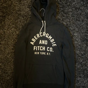 Svart hoodie Abercrombie & Fitch - Svart hoodie från Abercrombie & Fitch med vit broderad logga på bröstet och klassisk känguruficka. Tröjan har huva med vita snören och är långärmad. Perfekt för chill dagar eller när du vill ha en avslappnad look.