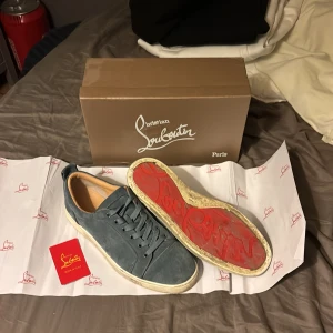 Blå mocka sneakers från Louboutin - Säljer ett par snygga blå sneakers från Christian Louboutin i mjuk mocka med klassisk röd sula och snörning. Skorna har en rund tå och vit platt sula, samt exklusiva detaljer som Louboutin-logga under sulan. Perfekt för dig som gillar lyxiga streetwear vibes.