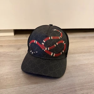 Svart Gucci keps med ormprint - Gucci keps i svart med klassiskt GG-mönster och en stor röd och vit orm på framsidan. Justerbar baktill med röd och svart detalj. Materialet är polyester och mesh, vilket gör den både snygg och luftig. Perfekt för dig som vill sticka ut med en lyxig streetstyle-accessoar. ÄKTA!! 