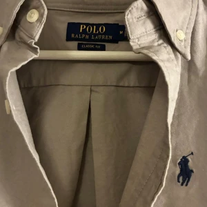 Grå skjorta från Polo Ralph Lauren - Snygg beige skjorta från Polo Ralph Lauren med klassisk passform och ikonisk blå broderad logga på bröstet. Skjortan har långa ärmar, knappar framtill och krage med knappdetaljer. Tillverkad i mjuk bomull som känns skön mot huden.