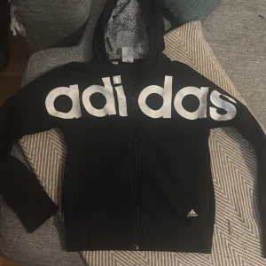 Svart hoodie från Adidas med dragkedja - Svart hoodie från Adidas med stor vit logga över bröstet och liten logga vid nederkant. Tröjan har huva, långa ärmar och hel dragkedja framtill. Perfekt för chill eller träning, med mjukt material och ribbade muddar.lite sliten i tyget se bild 3 endast där. 