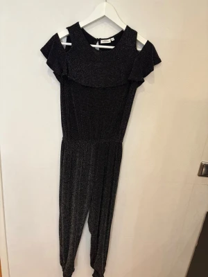 Glittrig svart jumpsuit med volang - Supersnygg svart jumpsuit från Kappahl med glittrigt tyg och volangdetalj över axlarna. Modellen har offshoulder och resår i midjan för en skön passform. Perfekt för dig som vill sticka ut med lite extra glitter och cool stil.