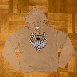 Beige Kenzo hoodie  - Säljer en beige hoodie från Kenzo i storlek S med ikoniskt tigerbroderi och Kenzo Paris-text på bröstet. Tröjan har huva med snörning, ribbade muddar och en klassisk känguruficka framtill. Perfekt för dig som gillar streetwear och snygga detaljer.