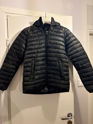 Svart pufferjacka från McKinley - Svart pufferjacka från McKinley med huva och dragkedja framtill. Jackan har horisontella quiltade sömmar, två fickor med dragkedja och en snygg, sportig look. Perfekt för kyliga dagar och enkel att matcha med din stil.