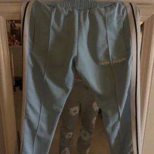 Palm Angels ljusblå mjukisbyxor - Snygga ljusblå mjukisbyxor från Palm Angels med vita och svarta sidoremsor. Byxorna har elastisk midja, raka ben och Palm Angels-logga på framsidan. Materialet är mjukt och har en lätt glansig finish som känns riktigt fräsch. Pris går att diskutera lite vid snabba affär