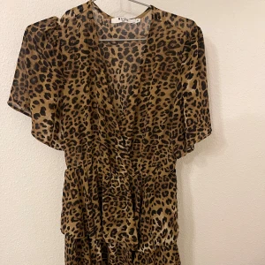 Leopardmönstrad kortklänning från NA-KD - Snygg kortklänning från NA-KD med leopardmönster i bruna och svarta toner. Klänningen har v-ringad hals, volangdetaljer och korta ärmar. Tillverkad i ett lätt och luftigt syntetmaterial som ger en skön känsla och cool look.