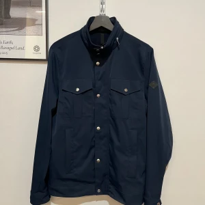 Mörkblå vindjacka från J.Lindeberg - Snygg mörkblå vindjacka från J.Lindeberg med hög krage, silverfärgade knappar och dragkedja. Jackan har två bröstfickor med lock, två sidofickor och ett diskret logomärke på ärmen. Perfekt för dig som gillar stilren och funktionell design.