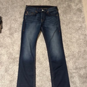 True Religion blå jeans, storlek 34 - Snygga blå jeans från True Religion med klassisk kontrastsöm och ikoniska broderade bakfickor. Modellen har rak passform och normal midja, tillverkade i slitstarkt denim med coola tvättade detaljer. Perfekt för dig som vill ha en streetig look.