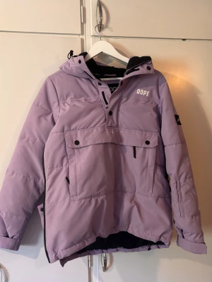 Snowboard Dope jacket  - Säljer en ljuslila pufferjacka från Dope med huva, stora fickor med knappar och dragkedja i sidan. Jackan har en cool streetstil och är perfekt för kalla dagar. Fodrad insida och snygga detaljer som logga på bröstet och ärmen.
