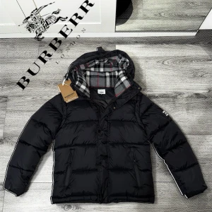 Svart Burberry pufferjacka  - Svart pufferjacka från Burberry med avtagbara ärmar, så du kan bära den som väst. Jackan har en huva med klassiskt rutigt Burberry-foder i grått, vitt och rött. Två dragkedjefickor fram och snygga detaljer på ärmen. Perfekt för kalla dagar och riktigt stilren.