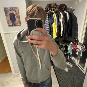 Grå hoodie från Polo Ralph Lauren - Säljer en grå hoodie från Polo Ralph Lauren med klassisk blå logga broderad på bröstet. Tröjan har huva med vita snören, magficka och ribbade muddar. Perfekt för chill dagar och enkel att matcha med jeans eller joggers. Kan gå ner i pris ifall snabbt köp 