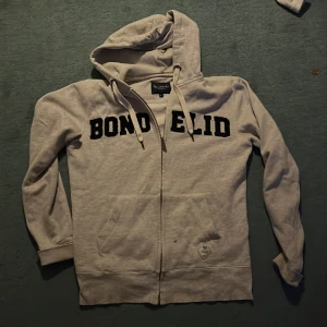 Beige hoodie från Bondelid med dragkedja - Säljer en beige hoodie från Bondelid i storlek S. Tröjan har svart text på bröstet, huva med snörning och praktisk dragkedja framtill. Mjuk bomullskänsla och fickor på magen gör den både chill och snygg. Perfekt för sköna dagar.