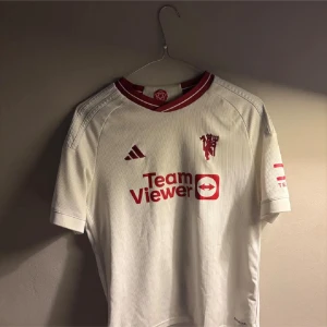 Manchester United vit matchtröja Adidas - Säljer en officiell Manchester United fotbollströja från Adidas i vit färg med röda detaljer. Tröjan har korta ärmar, ribbad struktur och klubbmärke på bröstet. Röd TeamViewer-logga framtill och DXC Technology-tryck på ärmen. Materialet är lätt och andas.