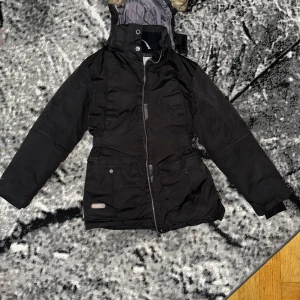 Svart vinterjacka från Protech - Svart vinterjacka från Protech med huva och fuskpälsdetalj. Jackan har dragkedja framtill, stora fickor med knappstängning och quiltad insida i grått. Perfekt för kalla dagar med sin varma och funktionella design.