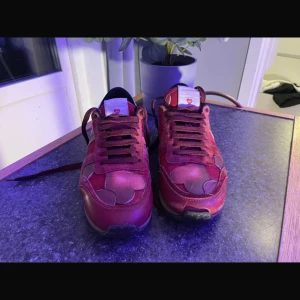 Valentino Rockrunner sneakers röd - Snygga Valentino Rockrunner sneakers i rött med hjärtan-mönster och detaljer i mocka och skinn. Skorna har snörning, platt sula och coola nitar på hälen. Perfekt för dig som vill sticka ut med ett unikt och trendigt par sneakers.