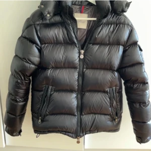 Moncler maya  - Säljer en svart pufferjacka från Moncler med huva och dragkedja framtill. Jackan har två fickor med dragkedja och en extra ficka med Moncler-logga på ärmen. Den är quiltad och har en glansig finish som ger en riktigt clean look.