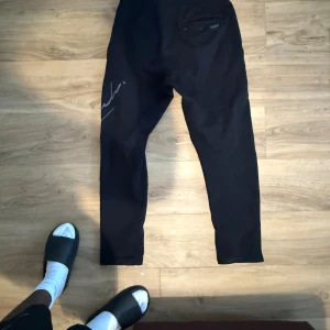 Svarta Hoodrich mjukisbyxor med tryck - Svarta mjukisbyxor från Hoodrich med tryck och logga på benet samt snörning i midjan. Byxorna har en slim passform och är tillverkade i mjukt bomullsmaterial. Perfekta för en chill och avslappnad stil.