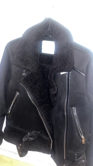 Svart stradivarius teddyjacka med bälte och dragkedja - Säljer en svart teddyjacka från märket Stradivarius med fluffig krage och foder, snygga detaljer i skinnimitation och två dragkedjefickor framtill. Jackan har bälte nedtill och grova dragkedjor som ger en cool vibe. Perfekt för dig som gillar streetstyle och vill hålla dig varm under exempelvis hösten.   Storlek: XS Nypris: 800 kr Mitt pris: 450 kr