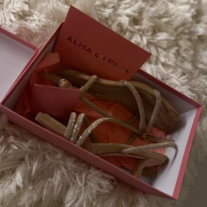 Beige klackskor med glittriga remmar - Snygga Alma en Pena-klackskor i beige mocka med glittriga remmar och fyrkantig tå. De har låg klack och spänne vid vristen. Perfekta för dig som gillar snygga detaljer och vill ha något extra till din outfit.