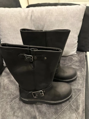 Svarta boots från Vagabond med spännen - Säljer ett par svarta boots från Vagabond i skinn med grov platt sula och två metallspännen. Modellen har rund tå och robust yttersula med tydligt mönster för extra grepp. Perfekt för dig som gillar chunky och stilrena skor.