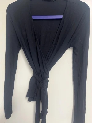 Svart omlottopp från Lager 157 - Svart omlottopp 'Lilja' från Lager 157 med långa ärmar och knyt i midjan. Toppen har v-ringad hals. 