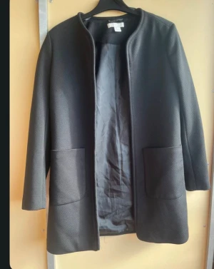 Svart rock med fickor från H&M - Säljer en stilren svart rock från H&M med två stora fickor framtill. Jackan har en enkel och rak design utan krage och är helsvart med lång ärm. Perfekt för dig som gillar minimalistisk stil och vill ha något som funkar till många outfits. Storlek M/L
