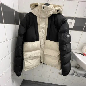 Svart och beige pufferjacka med huva - Säljer en fet pufferjacka från Never One i svart och beige. Jackan har stor huva, två rejäla fickor framtill och cool colorblock-design. Perfekt för kalla dagar och har en riktigt oversized vibe. Stängs med dragkedja och tryckknappar.