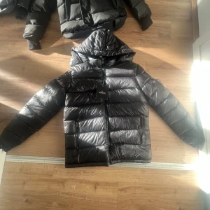 Svart Moncler pufferjacka med huva - Svart pufferjacka från Moncler med glansig finish, huva och dragkedja framtill. Jackan har Moncler-logga på ärmen, praktiska fickor med dragkedja och coolt serietidningsmönster på insidan. Perfekt för dig som vill ha en snygg och varm jacka med street vibe.