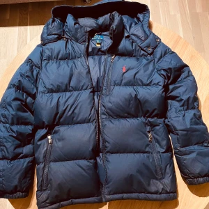 Mörkblå pufferjacka Polo Ralph Lauren XL. Juniorstorlek.  - Säljer en mörkblå pufferjacka från Polo Ralph Lauren i storlek XL(junior). Jackan har huva, två fickor med dragkedja och det klassiska röda Polo-logot broderat på bröstet. Perfekt för kalla dagar. Lite sliten i sömmen på vänster arm. I övrigt i mycket bra skick. 