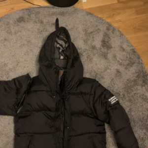 Svart pufferjacka från Burberry - Säljer en svart pufferjacka från Burberry med huva och klassiskt rutigt foder i grått, vitt och rött. Jackan har Burberry England-logga på ärmen och dragkedja framtill. Perfekt för kalla dagar och har en riktigt schysst streetstil. Problemet med jackan är att på ena ärmen går det inte att stänga men detta går och lösa hos en sybutik men då kanske man måste betala lite i mellan för att få detta problemet löst men annars är jackan bra kavlite men jackan går göra till en väst också.