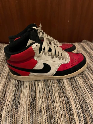 Nike Air Force 1 High röd/svart/vit - Säljer ett par Nike Air Force 1 High sneakers med klassisk färgblockning i rött, vitt och svart. Storlek: 42. Skorna har högt skaft, snörning och rund tå. Materialet är skinn med perforerade detaljer på tån och den ikoniska swoosh-loggan på sidan.